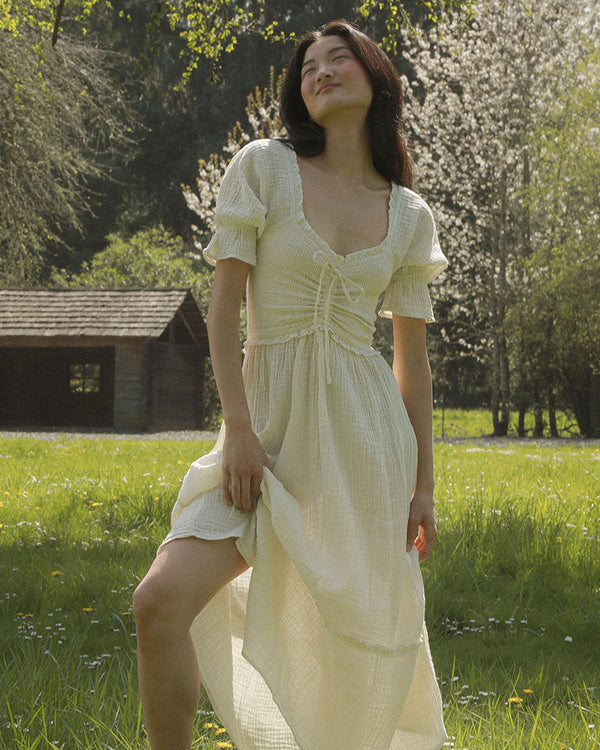 Woman in a white dress standing in a park with trees and a wooden structure in the background.
