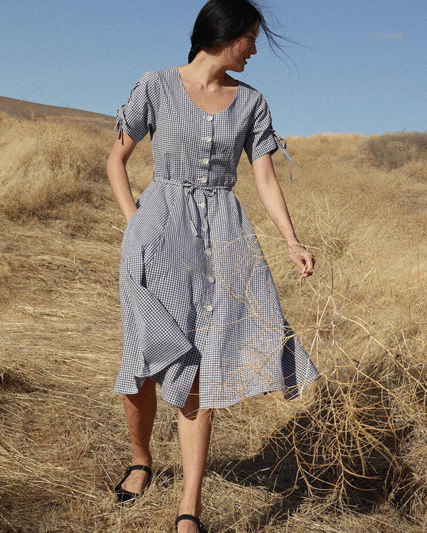 A woman standing in a dry grass field wearing a navy-blue cotton gingham check dress with a scoop neckline, mother-of-pearl buttons, and a self sash.