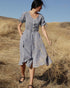 A woman standing in a dry grass field wearing a navy-blue cotton gingham check dress with a scoop neckline, mother-of-pearl buttons, and a self sash.