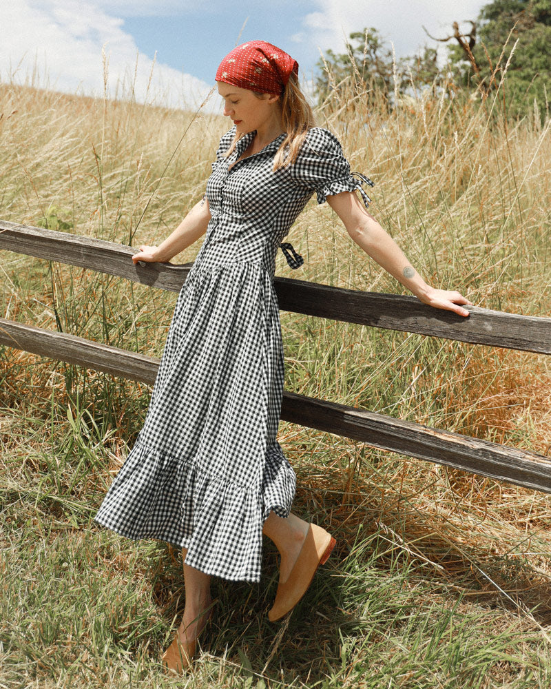 A woman wearing a black and white gingham midi dress with a red patterned headscarf stands outdoors leaning on a wooden fence in a grassy field.