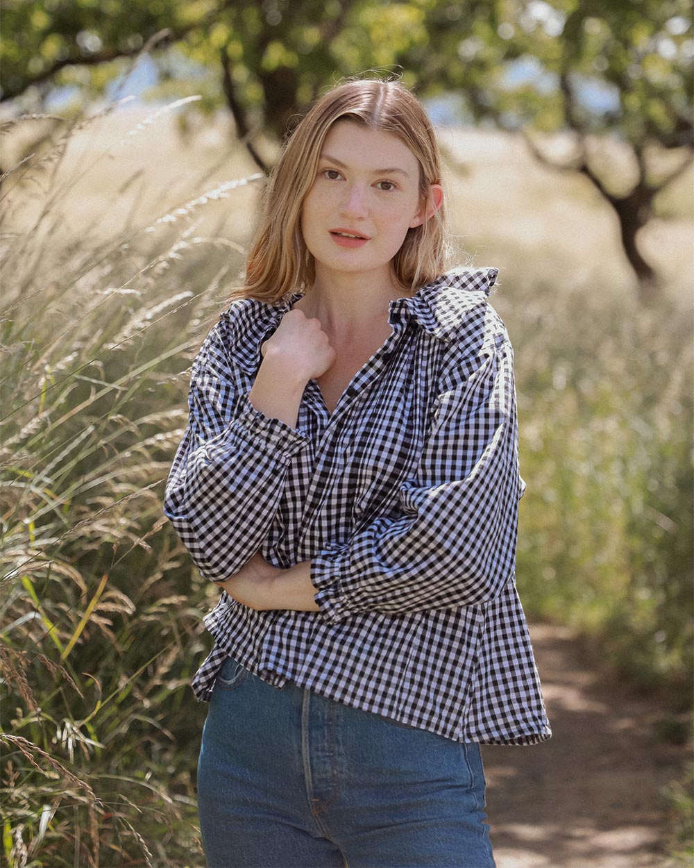 Woman wearing the Nesaea Top in black gingham with jeans standing in a field with tall grass and trees.