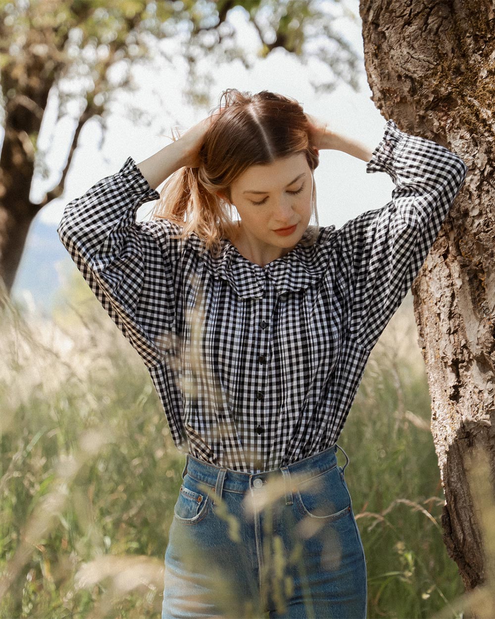 Woman in a checkered shirt and jeans standing in a natural setting with trees and grass.