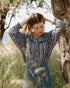 Woman in a checkered shirt and jeans standing in a natural setting with trees and grass.