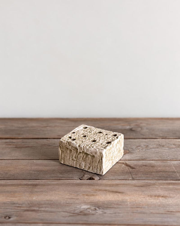 A small square decorative box with a textured, rustic stone-like finish and a lid featuring a grid of small holes, placed on a weathered wooden surface against a plain light gray wall.
